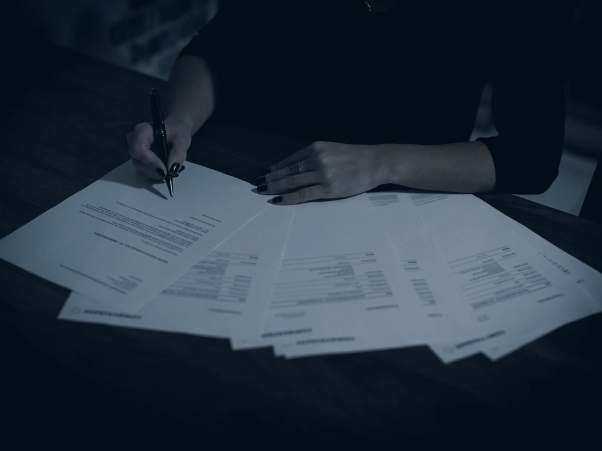 Documentos espalhados sobre uma mesa escura, com uma pessoa assinando